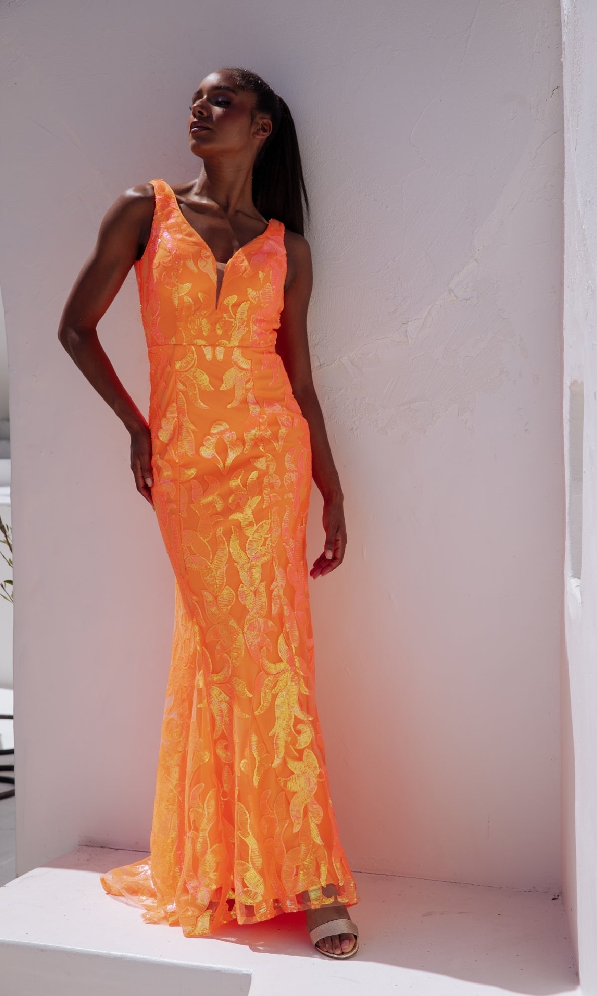 A woman poses by a white textured wall in the V-Back Long Sequin-Print Prom Dress: Eureka EK101—a sleeveless, floor-length bright orange gown with a deep V neckline.